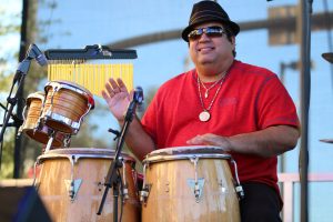 Drummer in red shirt, fedora and sunglasses playing the bongos during College Park JazzFest 2016